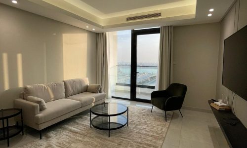 A modern AMWAJ flat for sale features a beige sofa, black armchair, round glass coffee table, large TV, and a sliding glass door leading to a balcony with a view. The ceiling has recessed lighting.