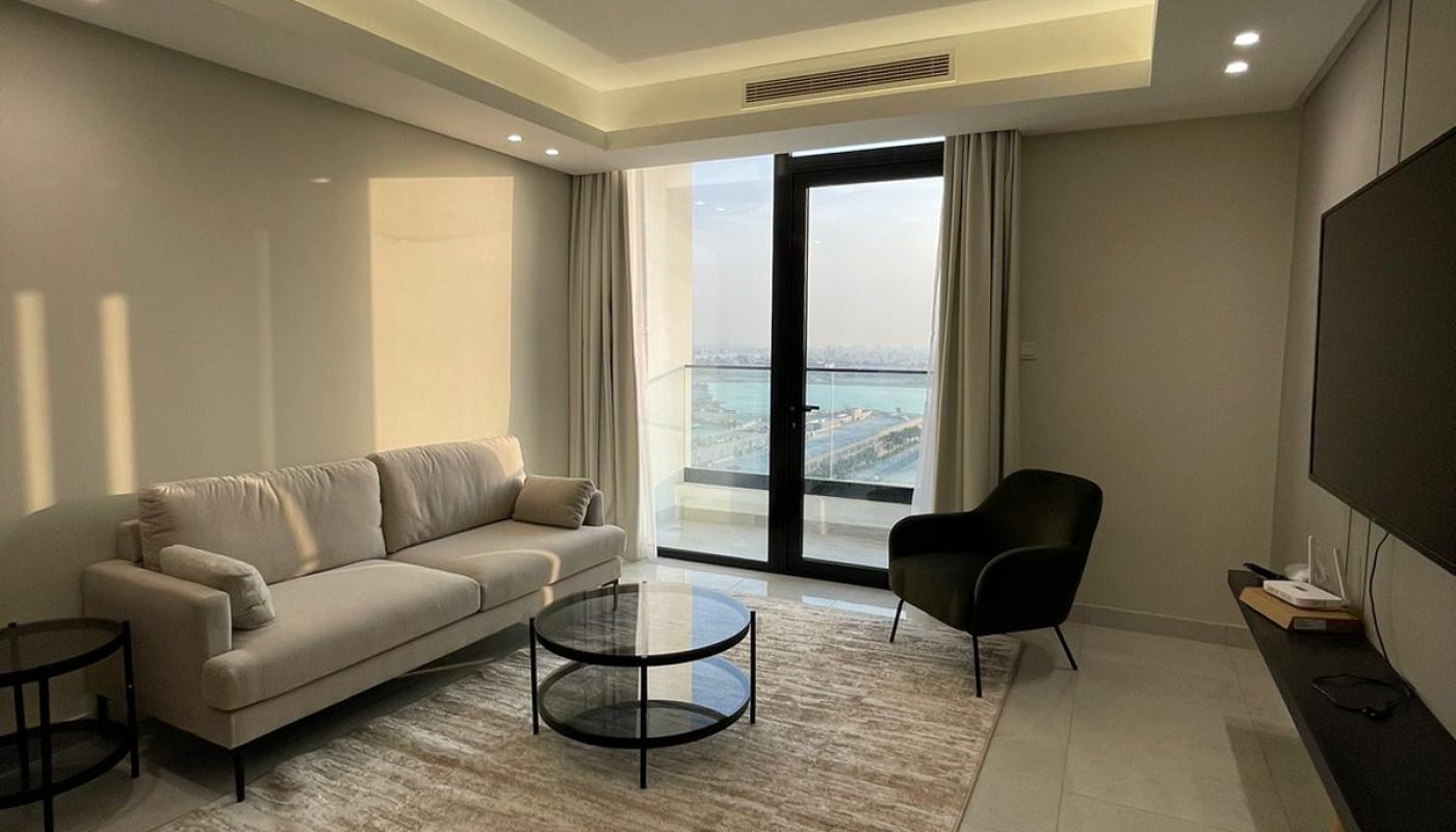 A modern living room with a beige sofa, black armchair, round glass coffee table, wall-mounted TV, large rug, and a glass door leading to a balcony with a view.