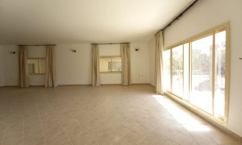 A spacious, empty room with tiled floors, large windows with beige curtains, and white walls lit by natural light.