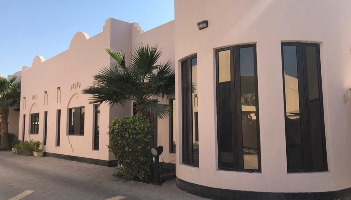 A modern, single-story building with a light exterior, large dark-tinted windows, palm trees, and paved walkway in front.