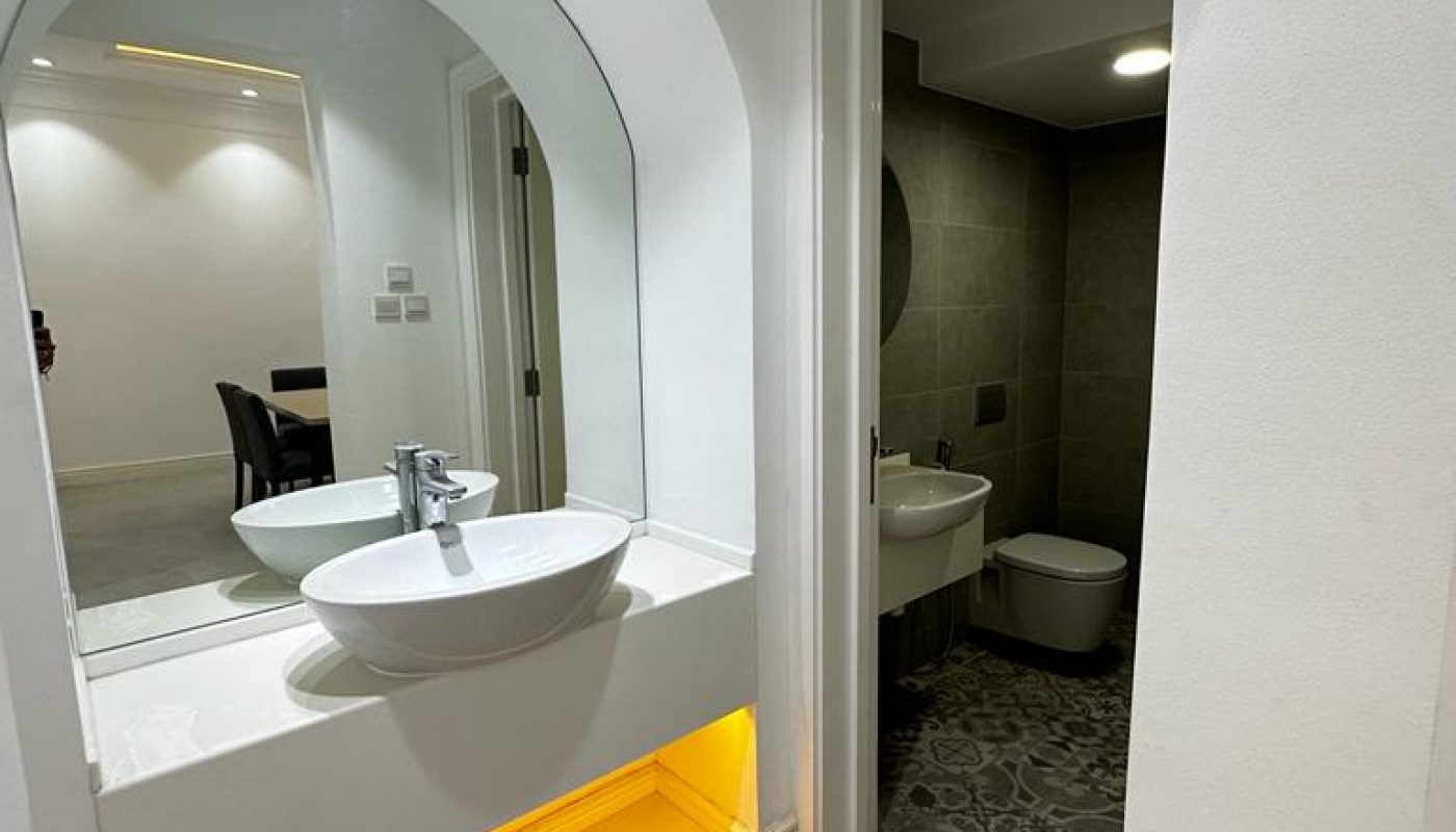 Modern bathroom with patterned floor tiles, a white sink on a counter with an arched mirror and yellow under-lighting, and a separate room for the toilet and bidet.