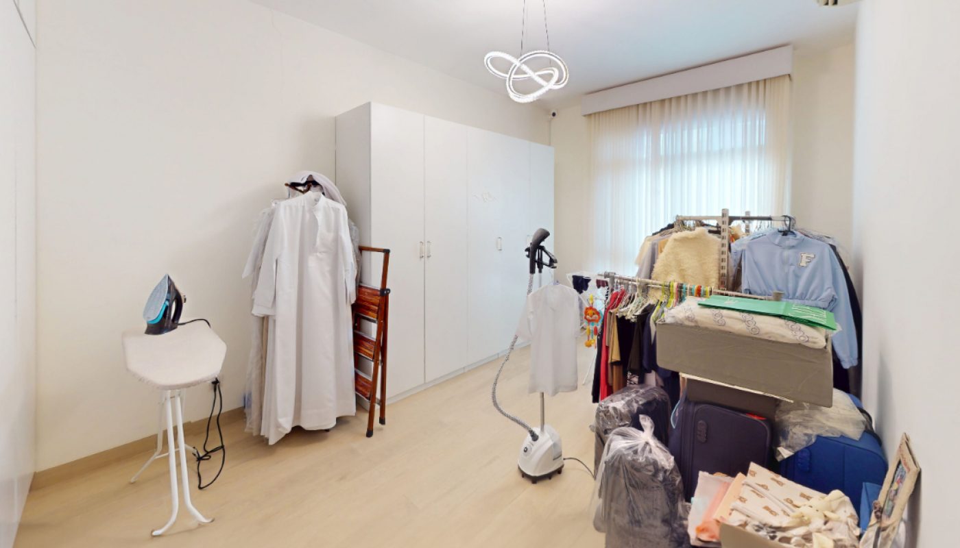 A tidy room with a wardrobe, ironing board, clothes steamer, hanging clothes, stacked boxes, and a window with sheer curtains.