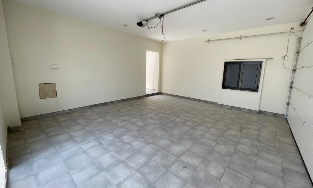 An empty garage with light-colored tiled floors, white walls, a window, and an open garage door leading outside. The space is lit by natural light streaming through the door. This elegant villa in Diyar Al Muharraq is now for sale.