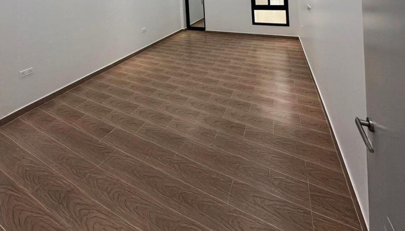Empty rectangular room with brown wood-patterned tile flooring, white walls, ceiling fan, recessed lighting, and a window next to a glass door at the far end.