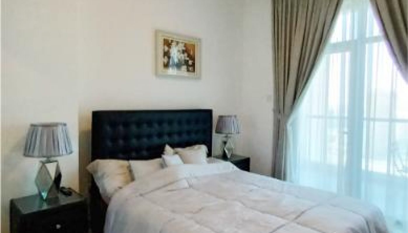A neatly made bed with a tufted headboard, two bedside tables with lamps, and large windows with curtains in a bright bedroom.