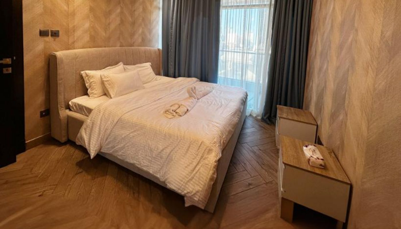 Modern bedroom with a double bed, white bedding, two nightstands, a ceiling light fixture, herringbone wood floor, and large window with curtains.