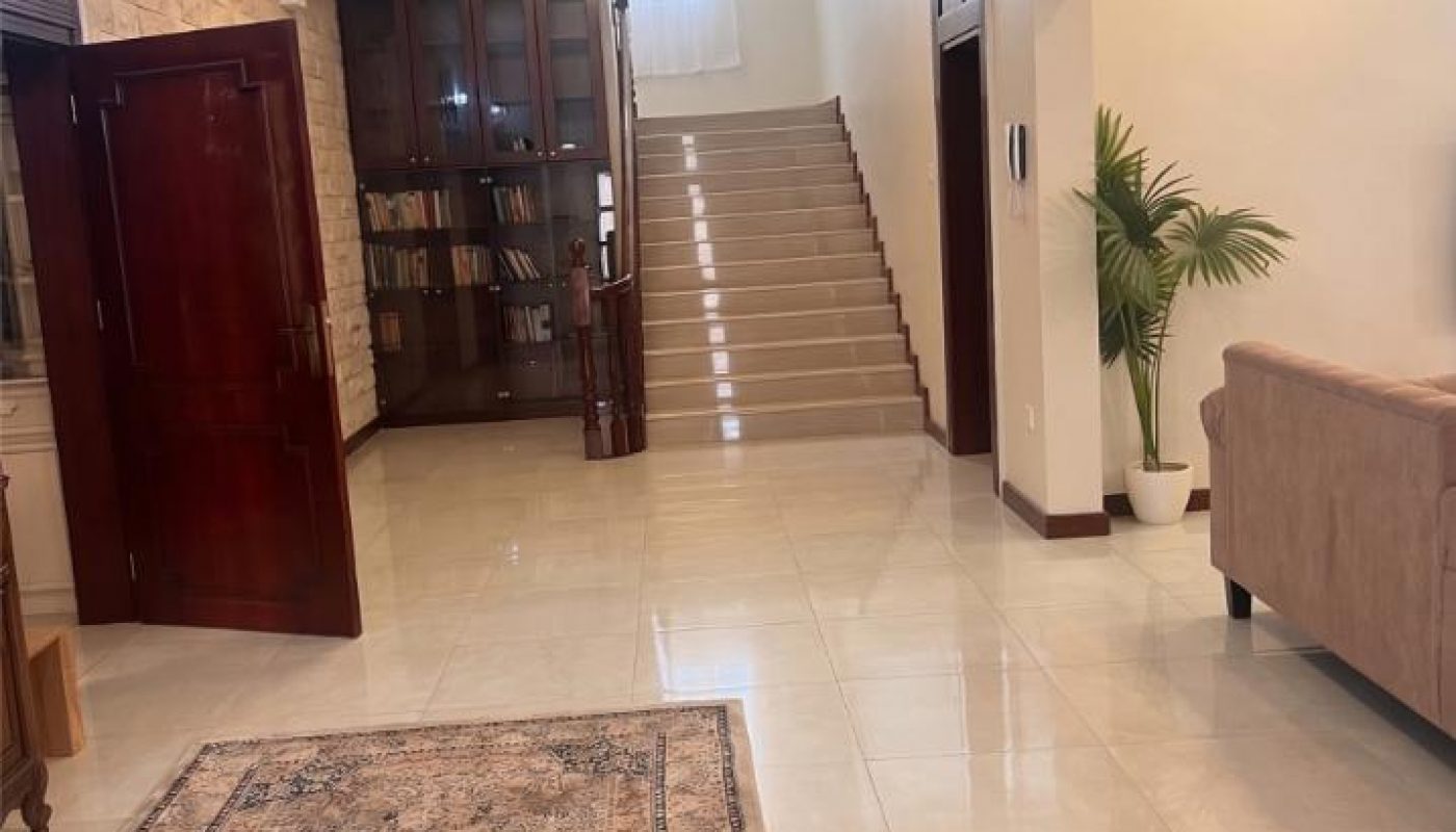 Spacious foyer with a patterned rug, tiled floor, beige sofa, indoor plant, wooden doors, bookshelf, and staircase leading to an upper floor.