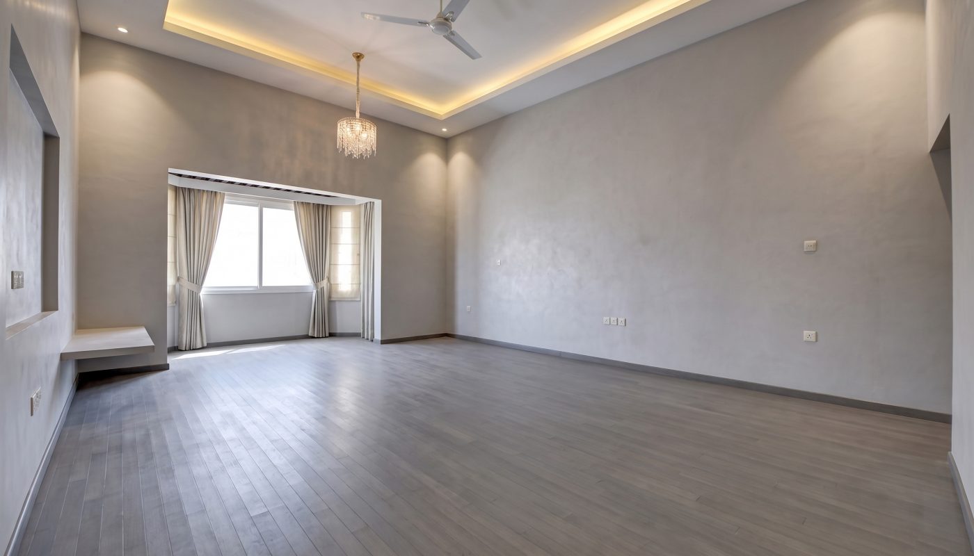 Spacious empty room with light gray wood floors, recessed ceiling lights, a ceiling fan, chandelier, large window with curtains, and plain walls.