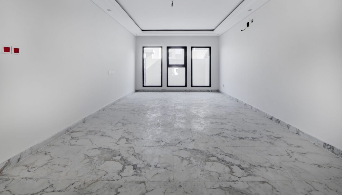 Empty room with white walls, a marble floor, three windows at the far end, and minimal fixtures on the walls and ceiling.