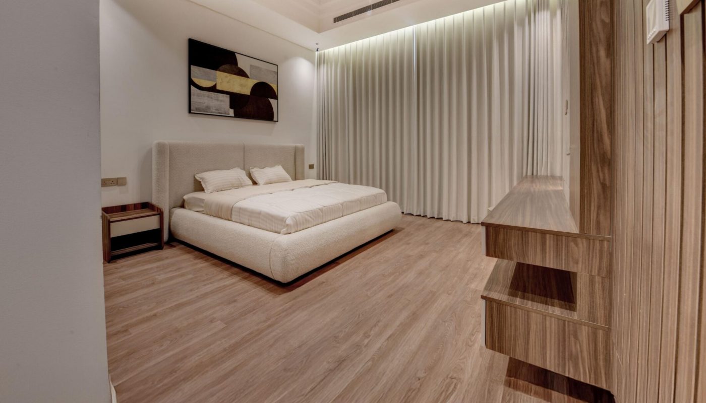 Minimalist bedroom with a low platform bed, neutral bedding, wooden floor, built-in wooden shelves, and a large curtain covering one wall. Abstract art hangs above the bed.
