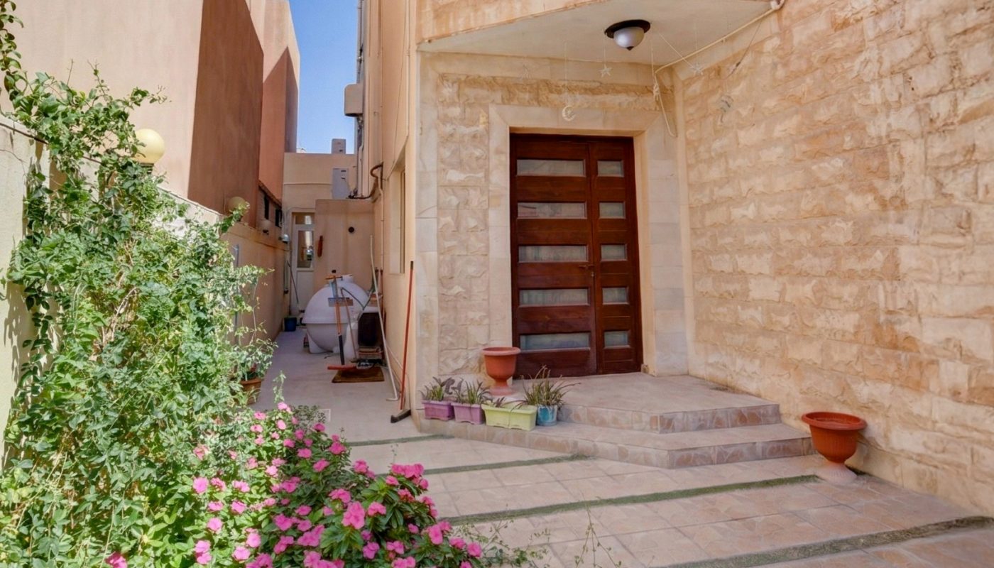 A stone building with a dark wooden door, tiled pathway, pink flowers in the foreground, and potted plants along the entrance under a clear blue sky.