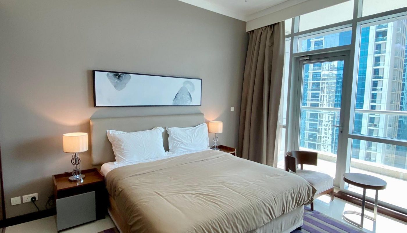 Modern bedroom with a large bed, two bedside tables with lamps, a striped rug, wall art, and a window with city view and balcony access.