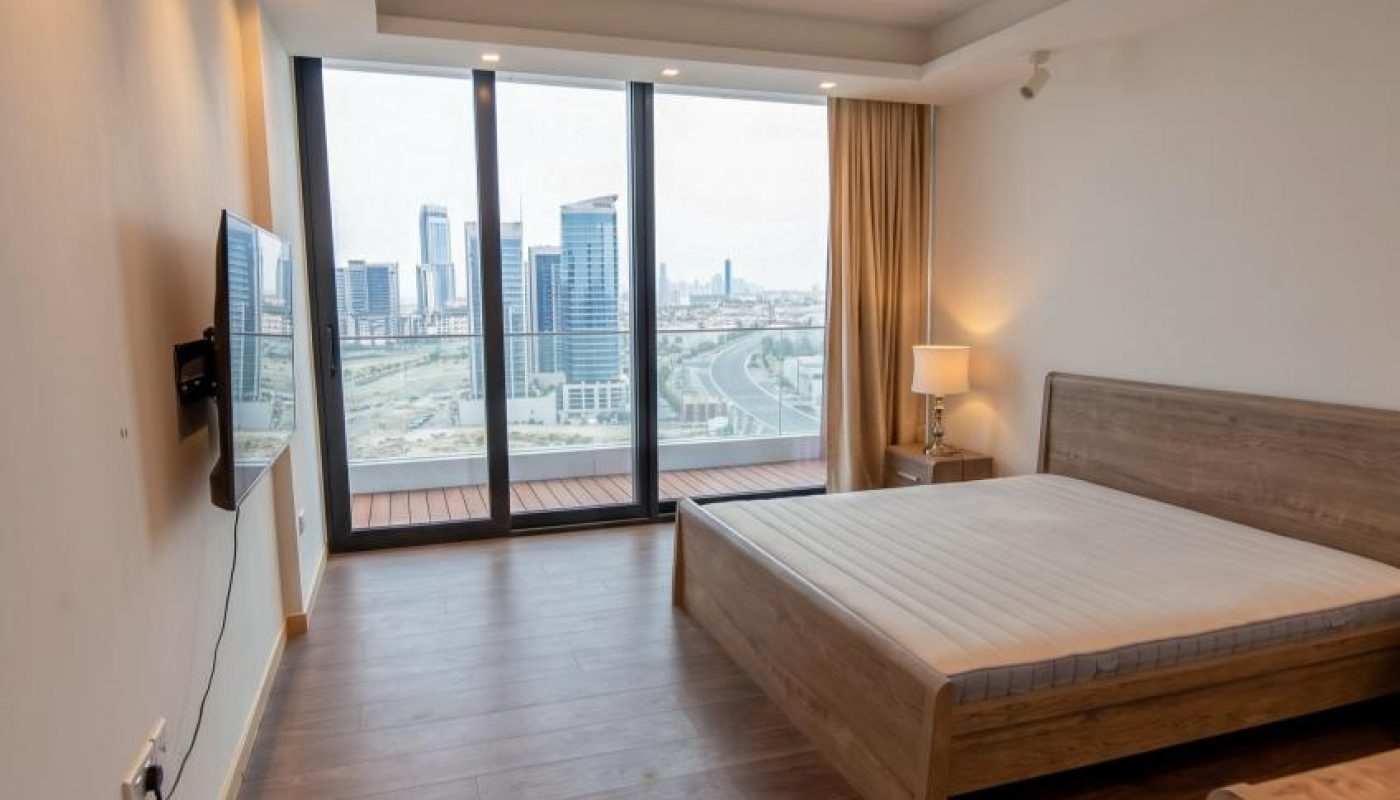 A modern bedroom with wooden flooring, a bed frame, wall-mounted TV, large floor-to-ceiling windows, and a city view in the background.