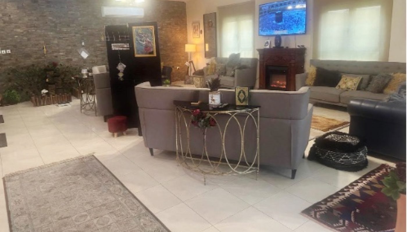 A spacious living room with a gray sectional sofa, wall-mounted TV, fireplace, light fixtures, and various decorative items, featuring neutral tones and brick accent wall.