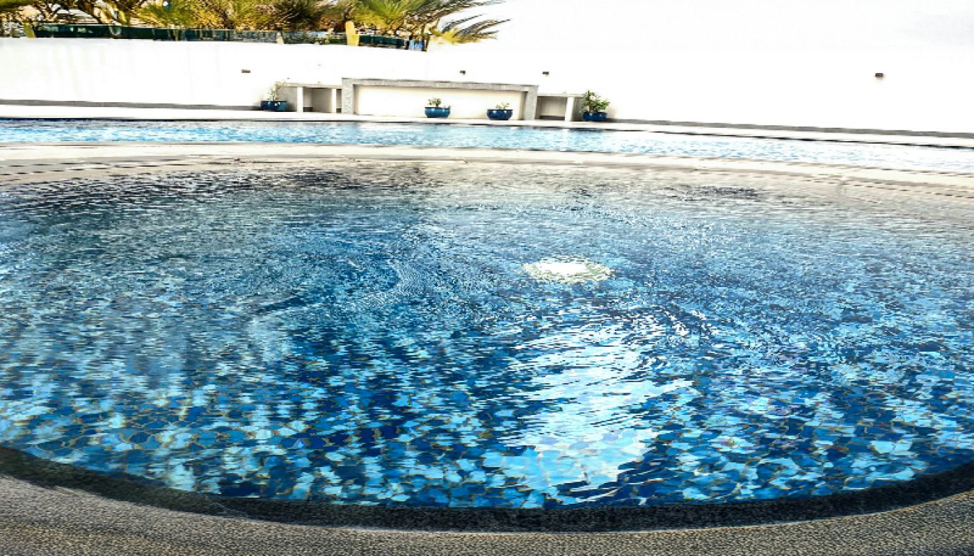 Outdoor round spa pool with bubbling water in the center, surrounded by gray tile, palm trees, and a white wall in the background.