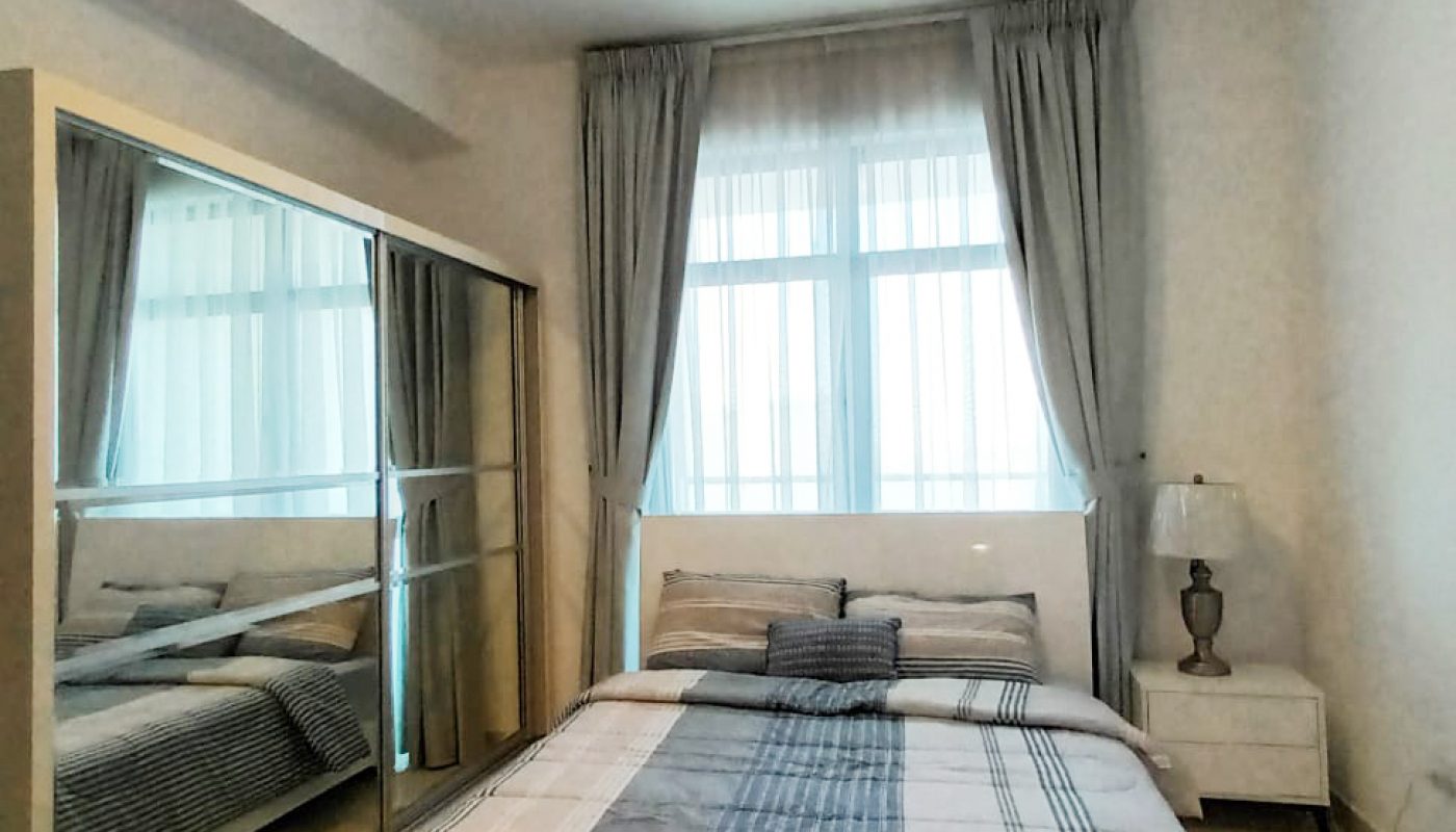 A neatly made bed with plaid bedding faces mirrored sliding closet doors in a bright bedroom with large window, light curtains, and a bedside table with a lamp.