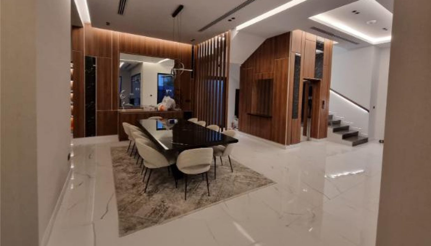 Modern dining area with a long black table, eight chairs, marble floor, wood panel walls, and stairs in the background.