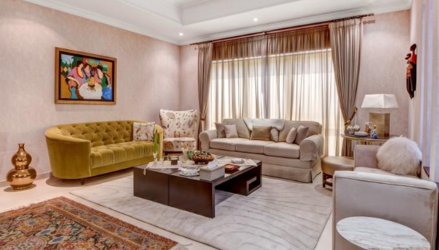 A spacious living room with two sofas, a coffee table, a patterned rug, large windows with curtains, artwork on the wall, and decorative accents on tables and in corners.