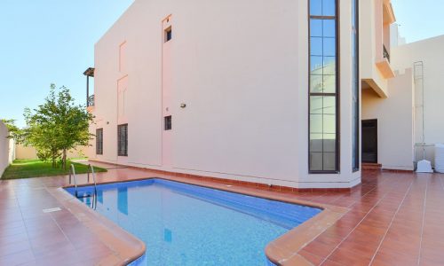 A modern 3BR villa in Al Areen boasts a two-story white structure with tall windows, a big garden, and a narrow, kidney-shaped outdoor pool lined with reddish tiles in the backyard.