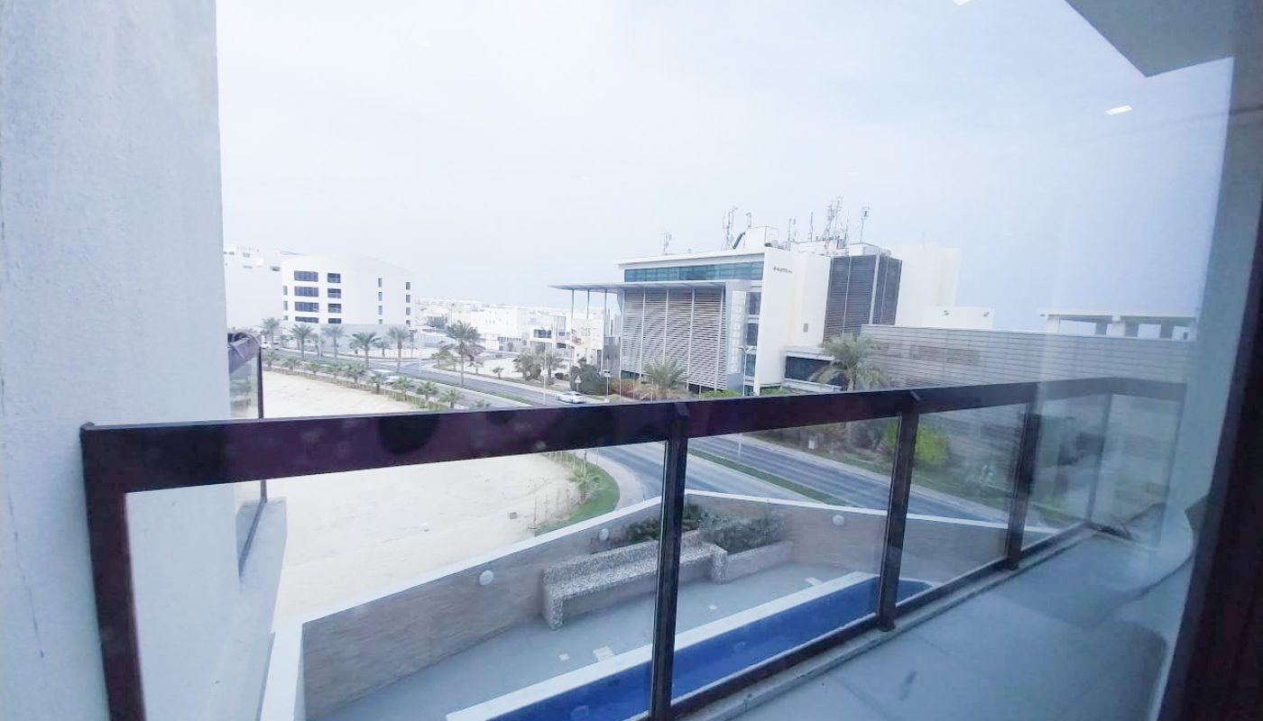 View from a balcony overlooking a modern building, glass railing, and a swimming pool, with other white buildings and a cloudy sky in the background.