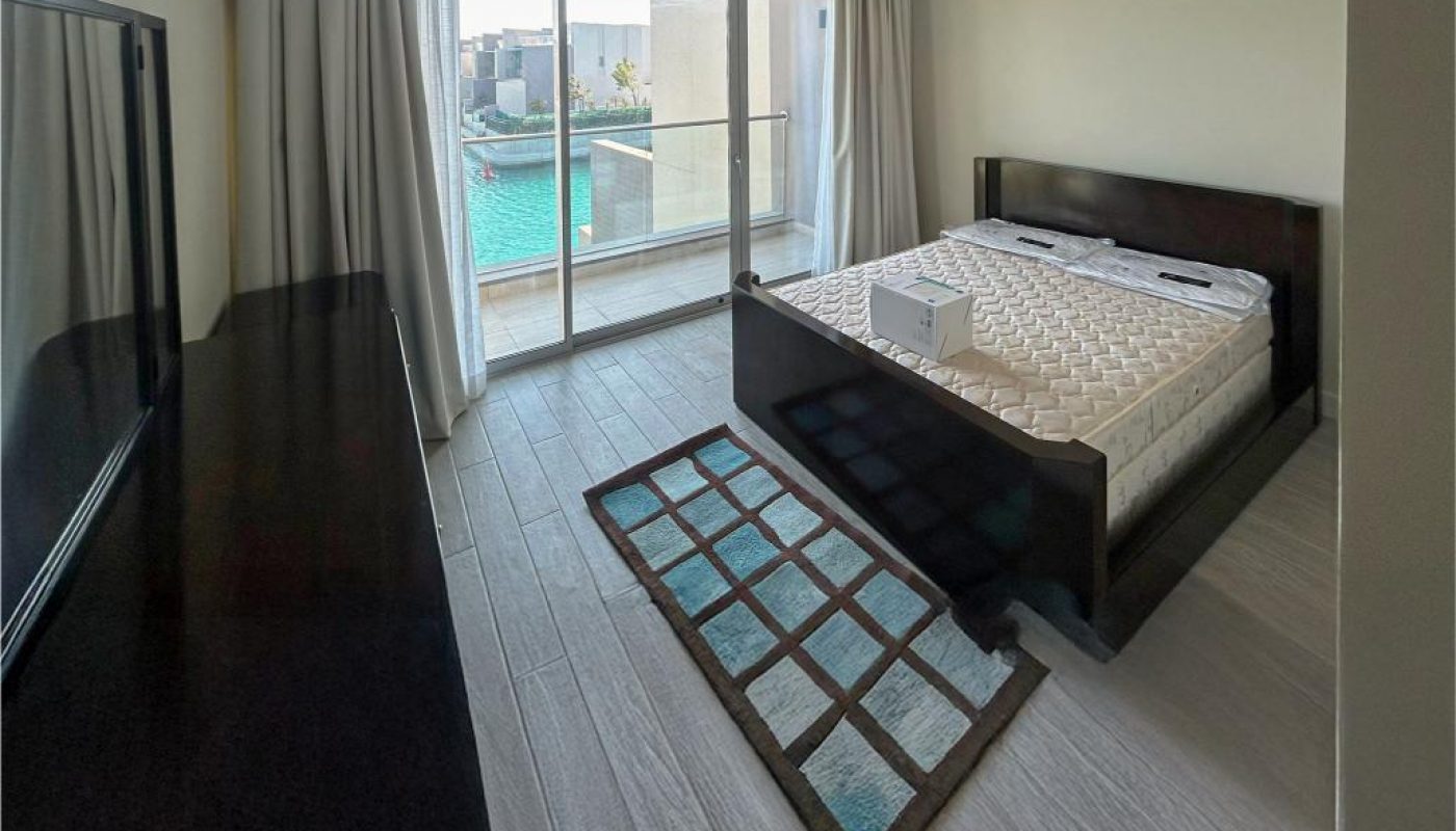 A modern bedroom with a large bed, wooden furniture, a TV, light gray flooring, and sliding glass doors leading to a balcony with a view of water and buildings outside.