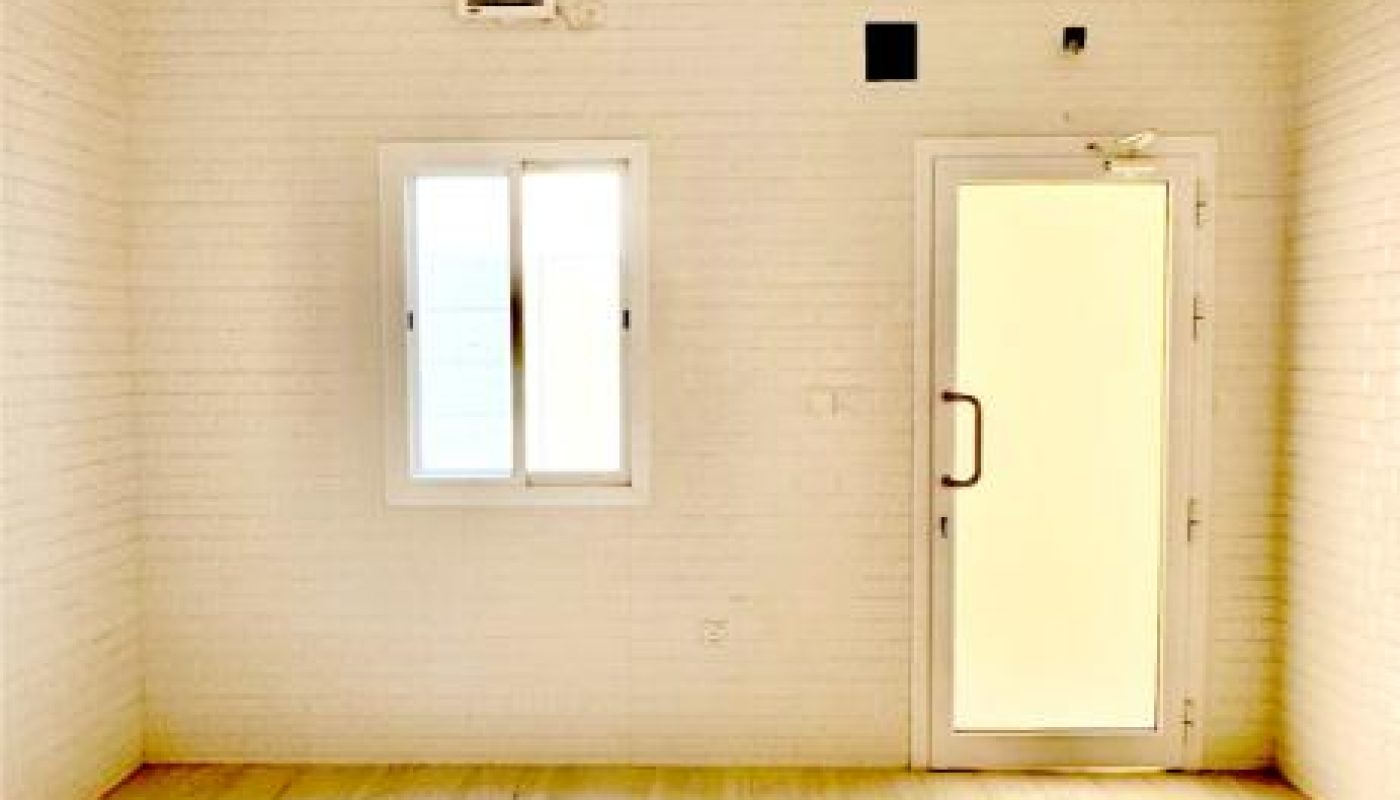 Empty room with light-colored tiled floor, white brick walls, a ceiling fan, a small window, and a glass door allowing natural light inside.