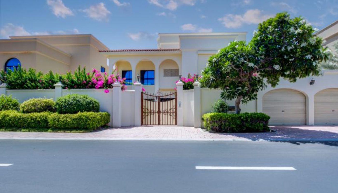 Two-story modern house with arched windows, a gated entrance, three garage doors, lush landscaping, and a clear blue sky overhead.