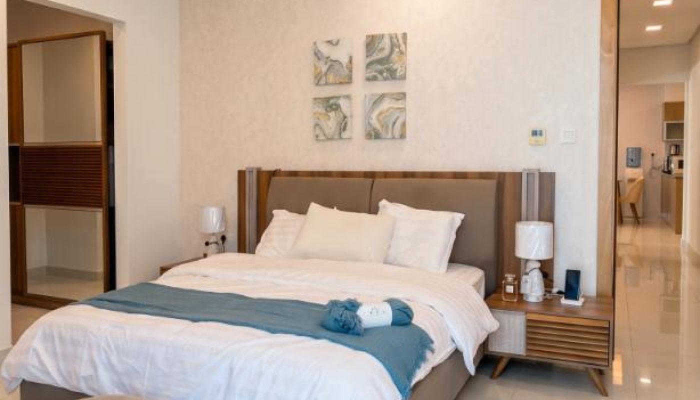 Modern bedroom with a double bed, white linens, wooden side tables, art on the wall, and a small beige stool on a glossy tile floor.