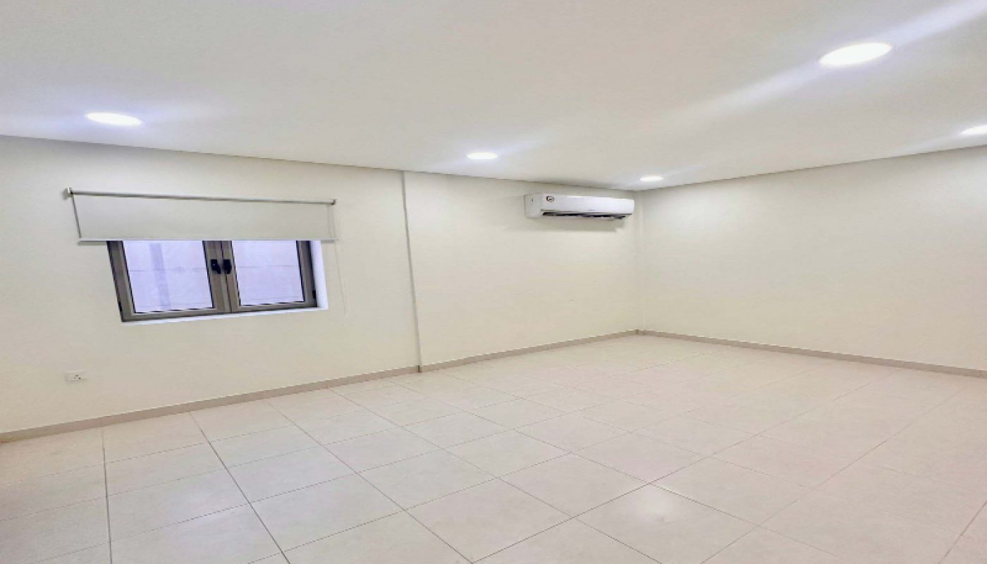 Empty room with cream-colored tiled floor, white walls, a window with a roller blind, ceiling lights, and a wall-mounted air conditioner.