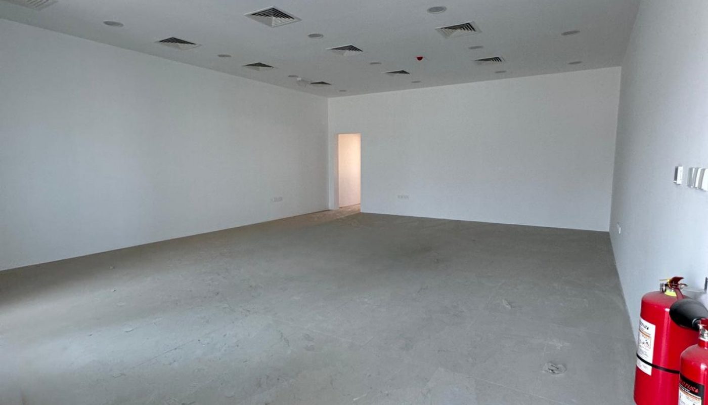 An empty, unfurnished white room with a concrete floor, ceiling vents, a doorway at the far end, and two fire extinguishers mounted on the right wall.