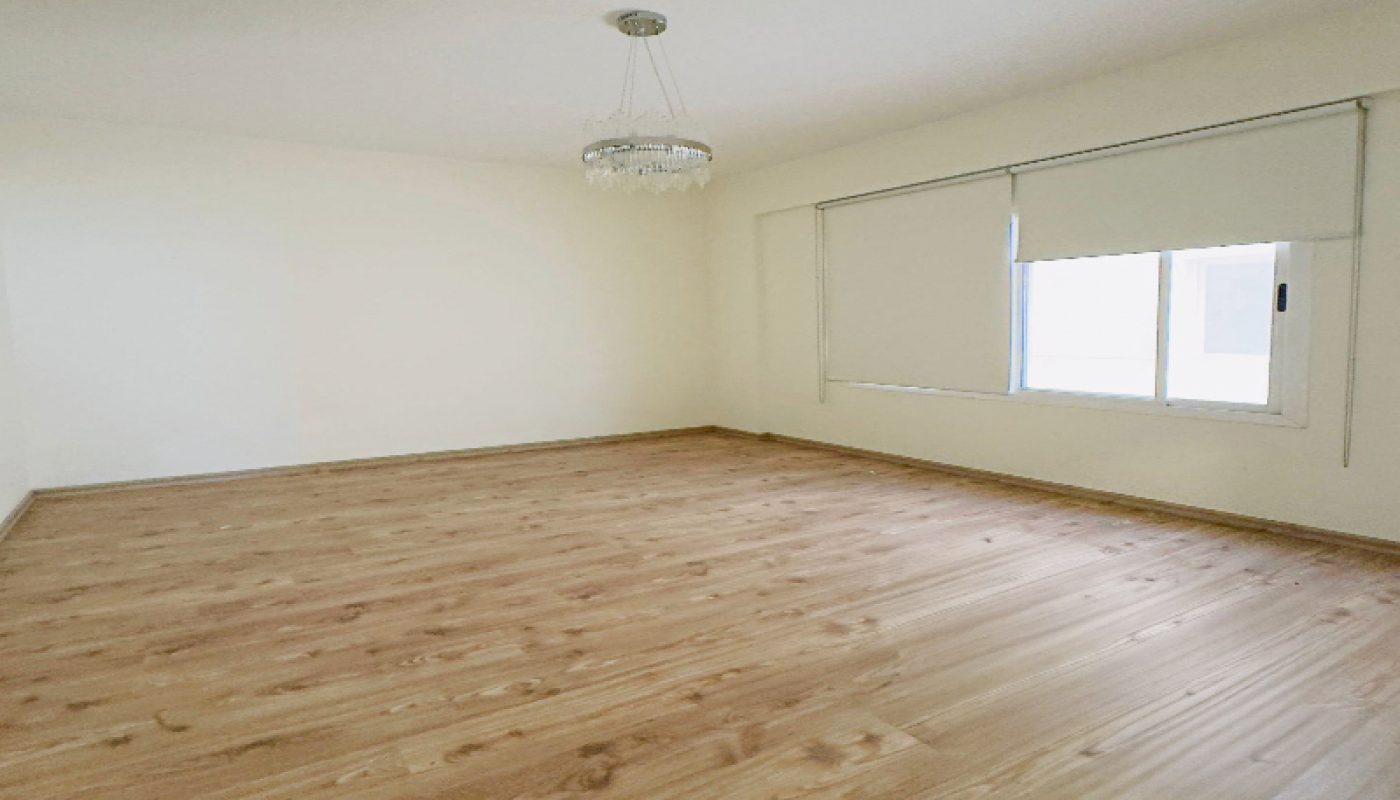 Empty room with light-colored wooden flooring, white walls, a ceiling light fixture, and a window with closed blinds.