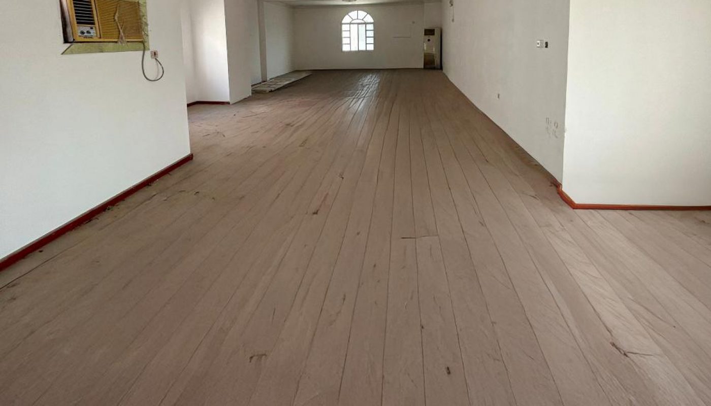 A spacious, empty room with light wood flooring, white walls, a large window at the far end, and a wall-mounted air conditioner.