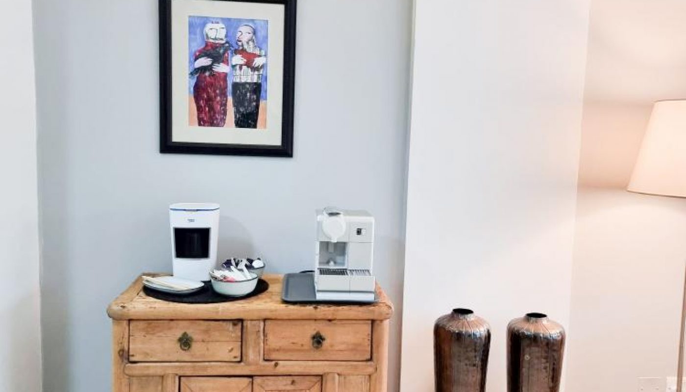 A wooden cabinet with two coffee machines and cups sits below a framed artwork and wall light; two tall vases and a floor lamp are on the right.