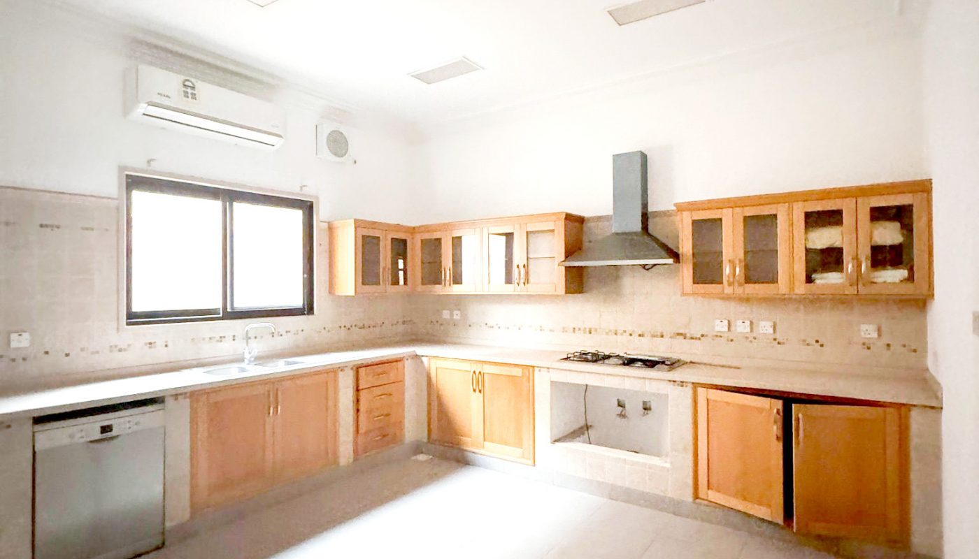 Spacious, empty kitchen with wooden cabinets, tiled floor, dishwasher, built-in oven, range hood, and large window letting in natural light.