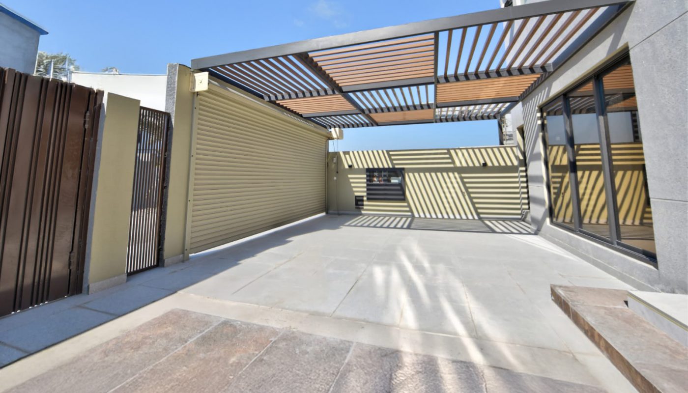 Modern outdoor patio with a slatted pergola attached to a contemporary building, casting striped shadows on the ground under a clear blue sky.