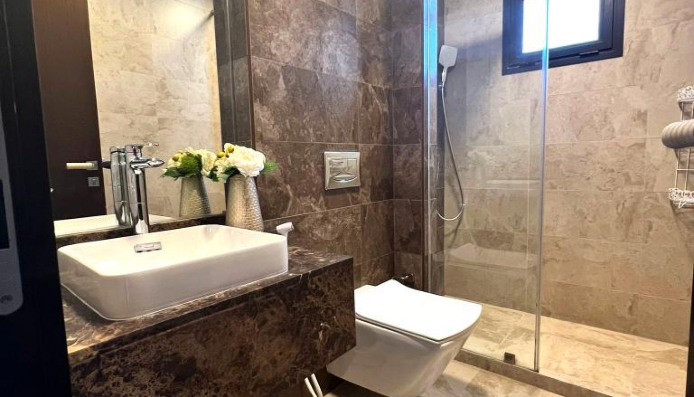 Modern bathroom with marble walls and floor, a wall-mounted toilet, glass-enclosed shower, white sink, flowers in a vase, and a trash bin under the counter.