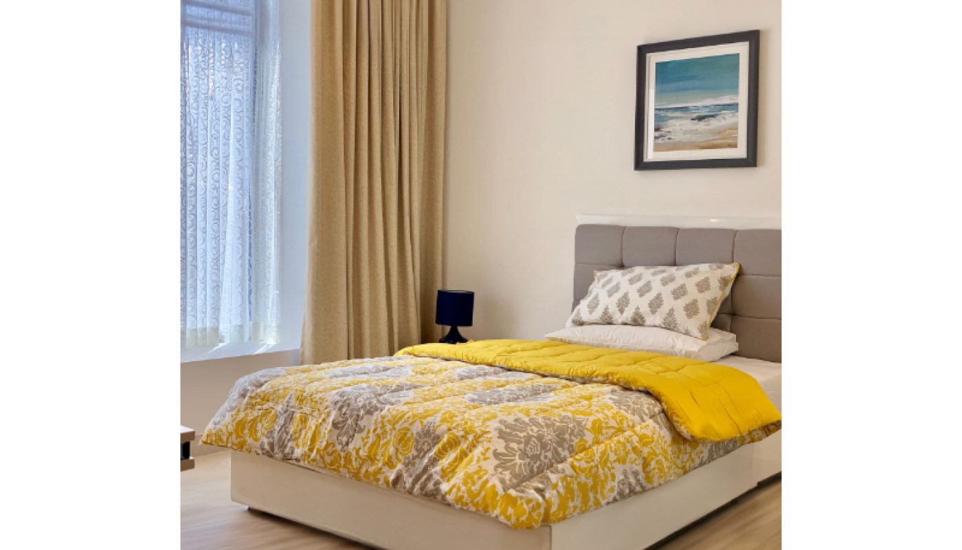 A neatly made bed with a yellow and gray comforter, patterned pillow, and beige curtains beside a window in a bright, modern bedroom.