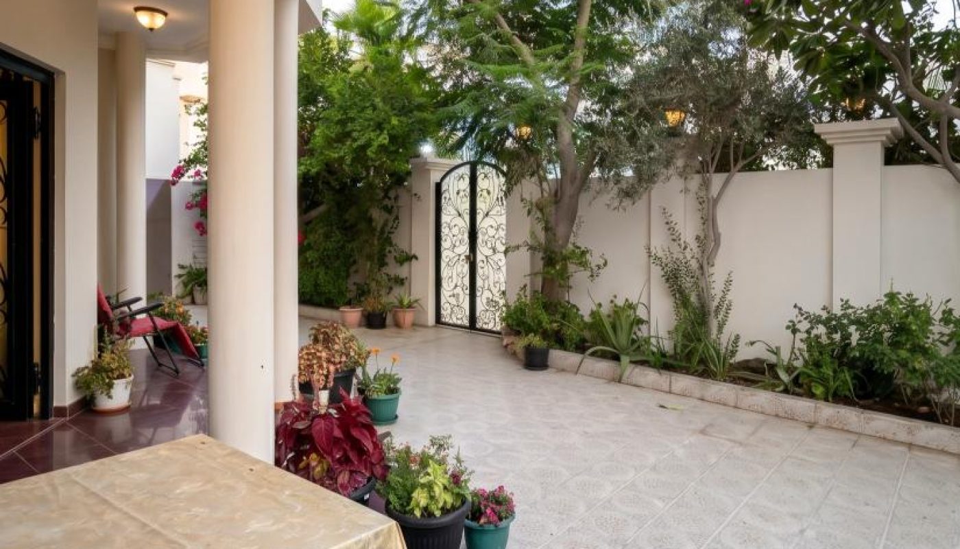 Covered patio with tiled floor, potted plants, a table with a gold-patterned cloth, chairs, and a white gate leading to a garden area surrounded by trees and shrubs.