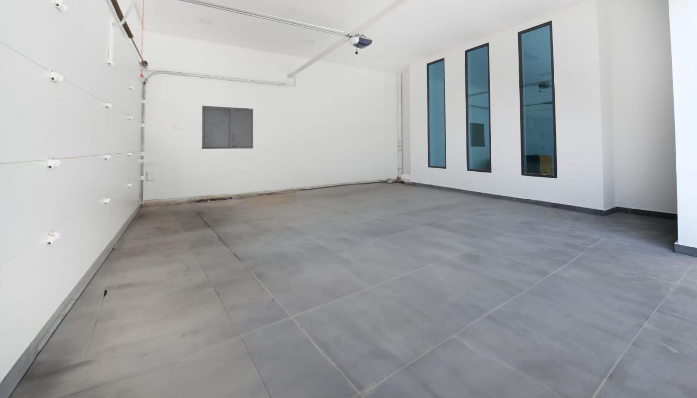 Empty modern garage with white walls, a gray tiled floor, large windows on the right, and an open garage door.