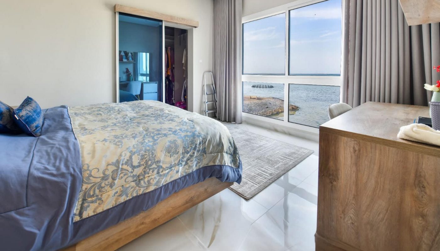 Bedroom with a large window view of the sea, a double bed with patterned bedding, a wooden dresser, and a shelf. Light-colored floor tiles and gray curtains complete the décor.