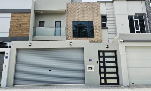 A luxurious modern two-story villa with a flat roof, large garage door, and a front door with glass panels. The exterior features varied textures including wood, stone, and metal. The front area is unpaved.