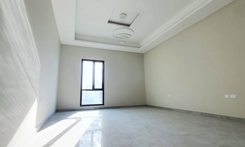 A spacious, empty room with light-colored walls, a large floor-to-ceiling window, and a chandelier in the center of the ceiling. The light gray tile floor reflects the natural light flooding in, imparting an almost auto-draft feel to this serene space.