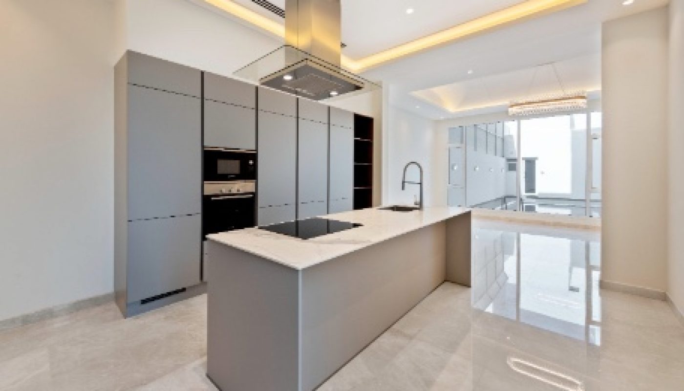 Modern kitchen with grey cabinetry, large island with built-in cooktop, overhead range hood, integrated appliances, and minimalist lighting in a bright, spacious room with marble flooring.