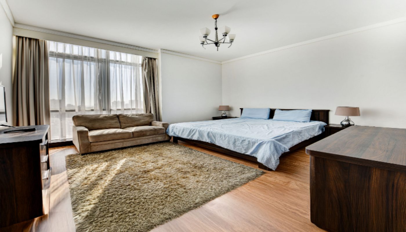 Spacious bedroom with a large bed, a brown sofa, wooden furniture, a beige rug, and a window with curtains letting in natural light.