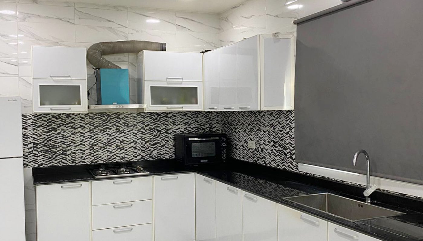 Modern kitchen with white cabinets, black countertops, a stainless steel sink, refrigerator, microwave, stove, and tiled floor and walls. Ceiling fan and roller blind are also visible.