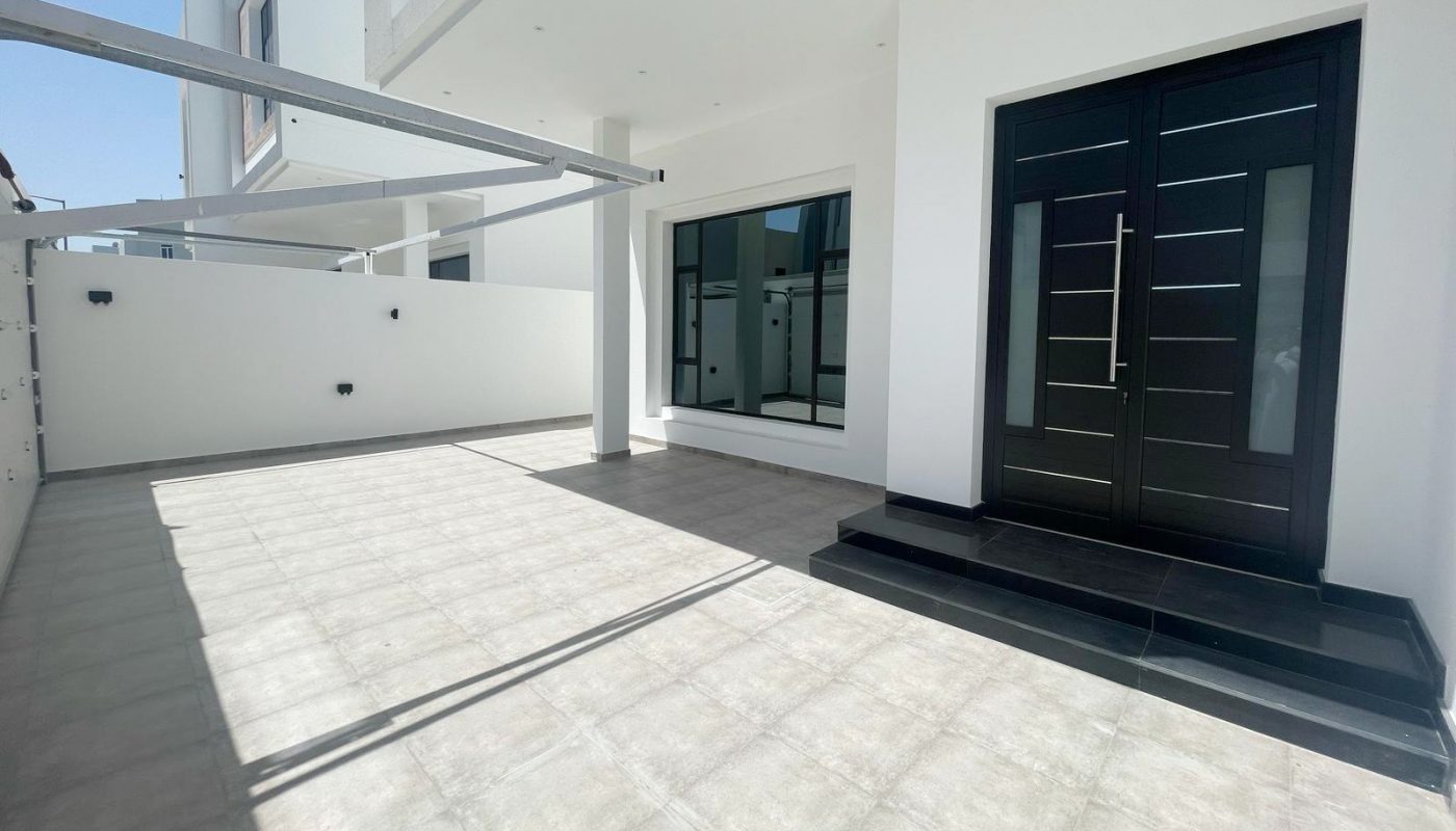 Spacious, modern house exterior with large tiled patio, black double doors, white walls, and a covered area on a sunny day.