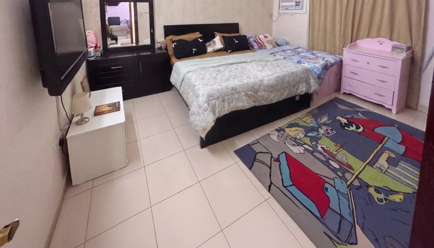 A bedroom with a large bed, TV, dresser, pink chest of drawers, air conditioner, and a colorful cartoon rug on tiled flooring.
