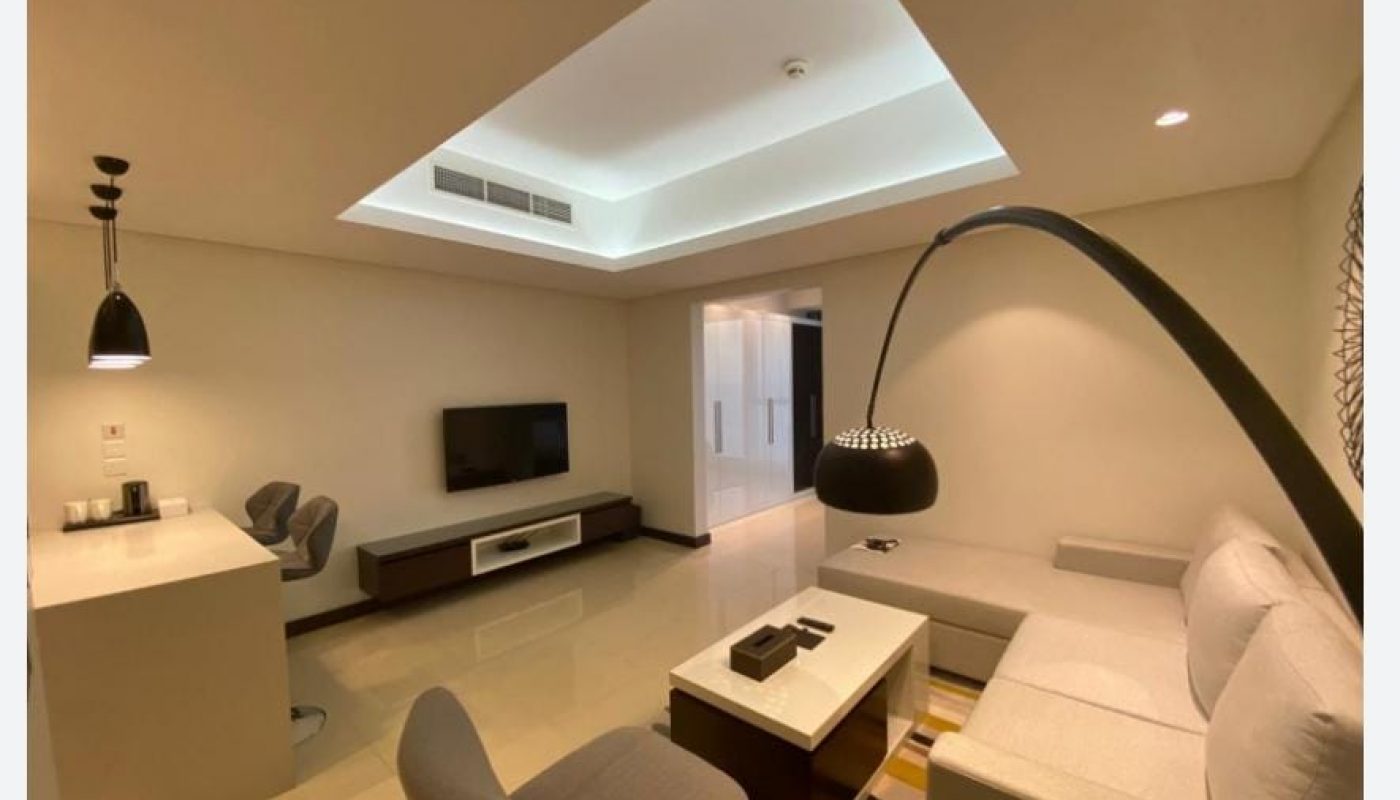 Modern living room with beige walls, a sectional sofa, a flat-screen TV on a low stand, a coffee table, and a kitchen counter with stools; ceiling has recessed lighting.