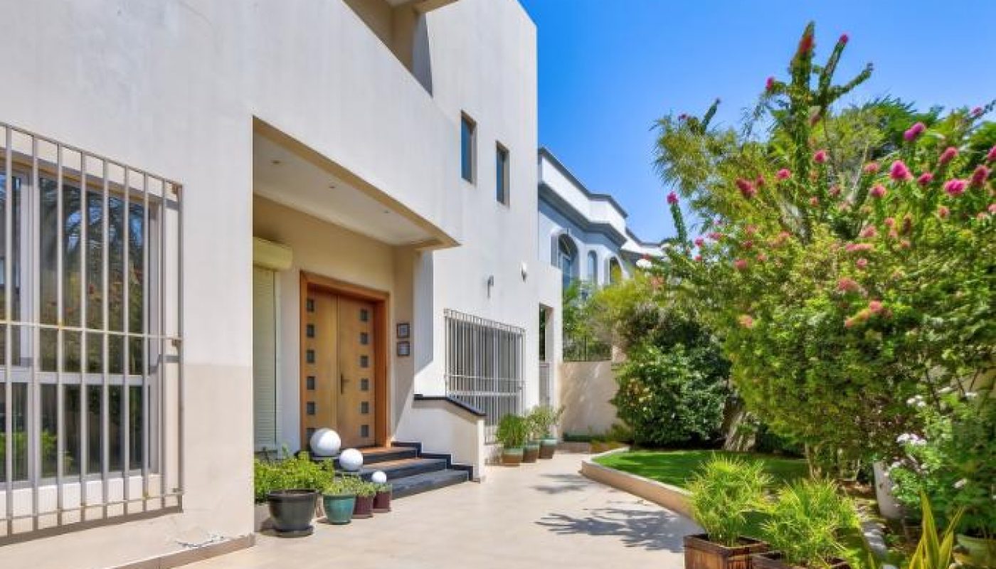 Modern house exterior with beige walls, large windows, a wooden front door, tiled patio, potted plants, and a green garden on a sunny day.