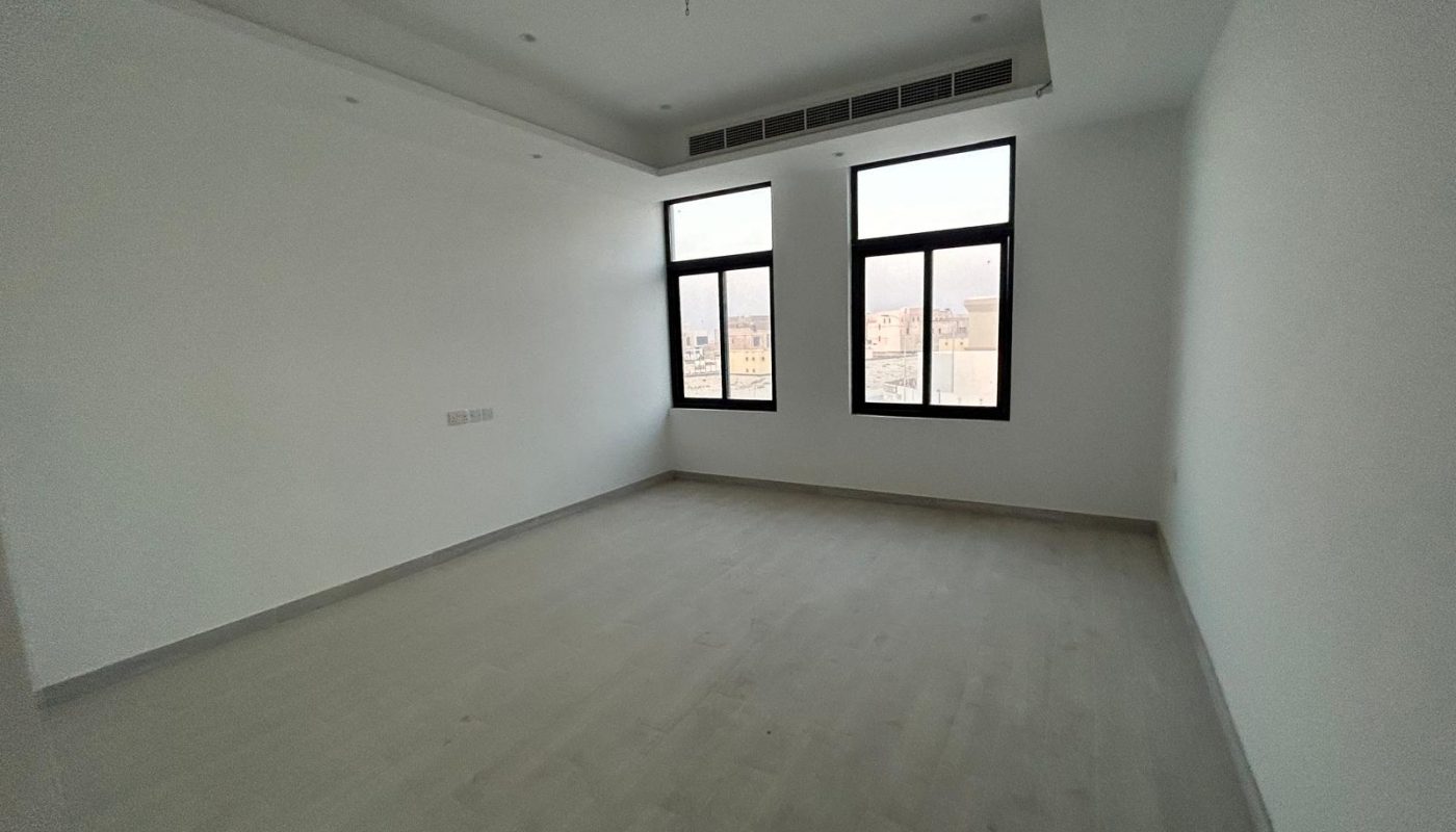 Empty room with white walls, light wood floor, two large windows, recessed ceiling, and air conditioning vents above the windows.
